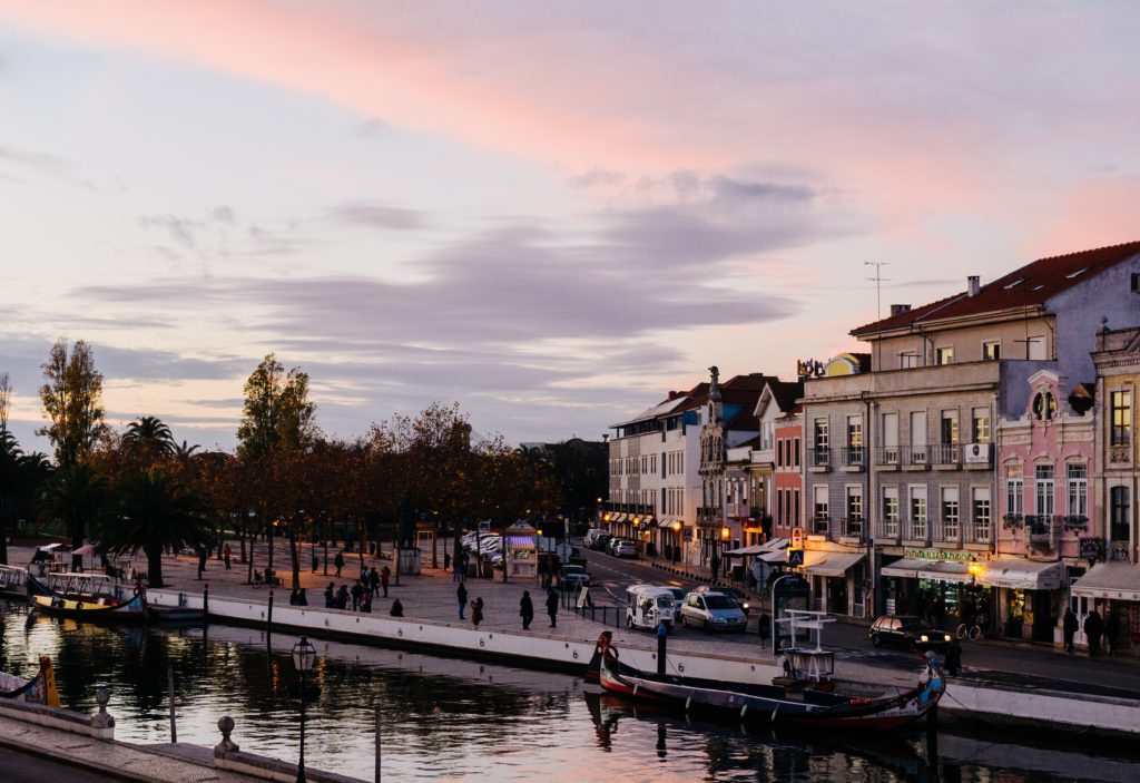 Vista de Aveiro ao entardecer, com a ria calma, um barco moliceiro parado e as casinhas típicas nas margens.