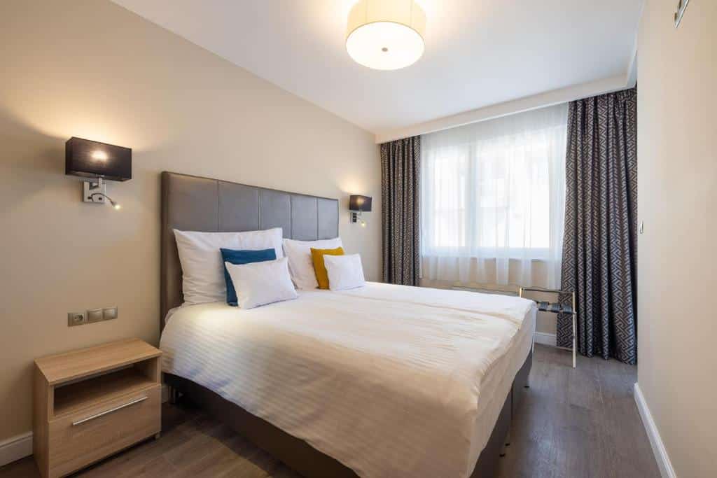 Uma cama bem arrumada do Silver Crown Hotel &amp; Residence, com roupa de cama branca, dois travesseiros de destaque e uma mesa de cabeceira, num quarto moderno e bem iluminado.