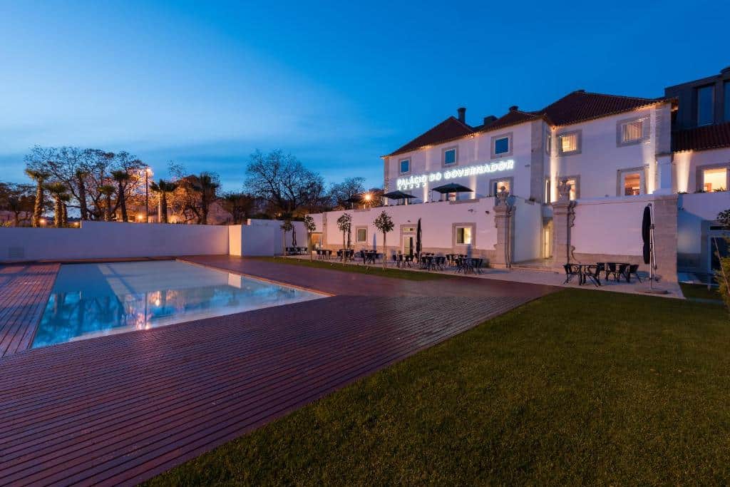 Piscina exterior moderna com deck de madeira junto a um grande edifício branco ao anoitecer, com zonas de estar e iluminação, no Palácio do Governador.