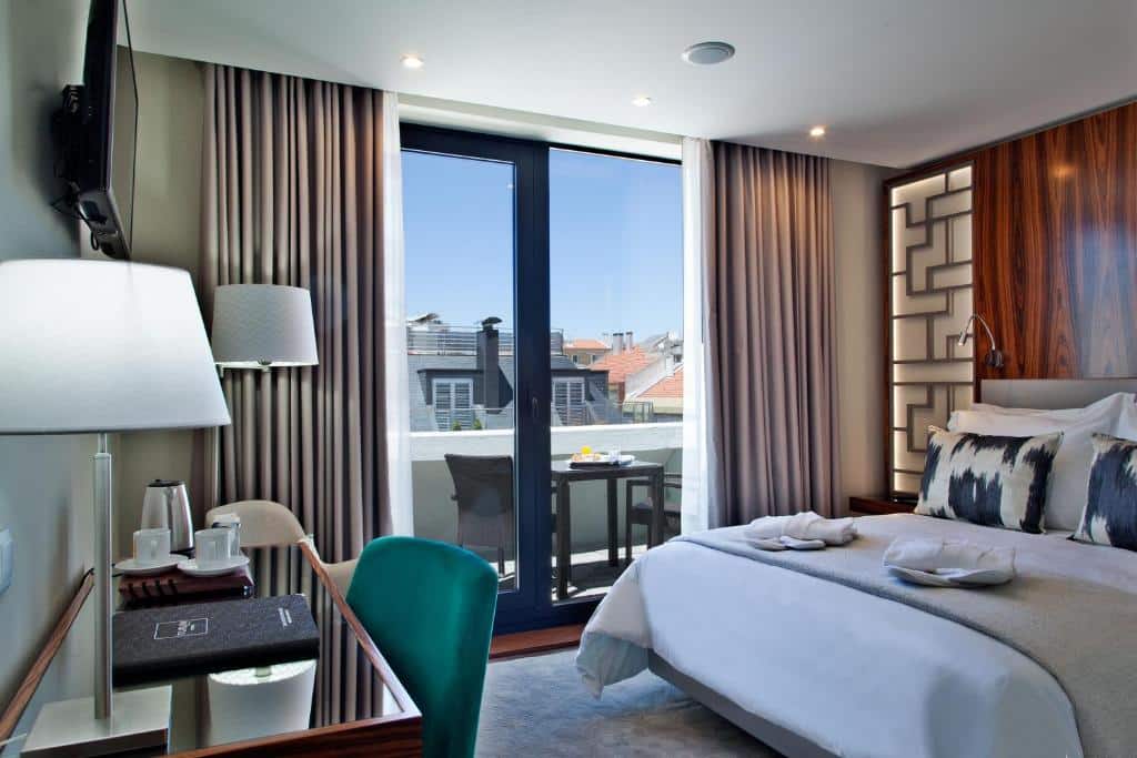 Quarto do TURIM Saldanha Hotel com uma cama de casal, uma varanda com cortinas e mesa com duas cadeiras do lado esquerdo e, em frente à cama, uma televisão e uma secretária com poltrona.