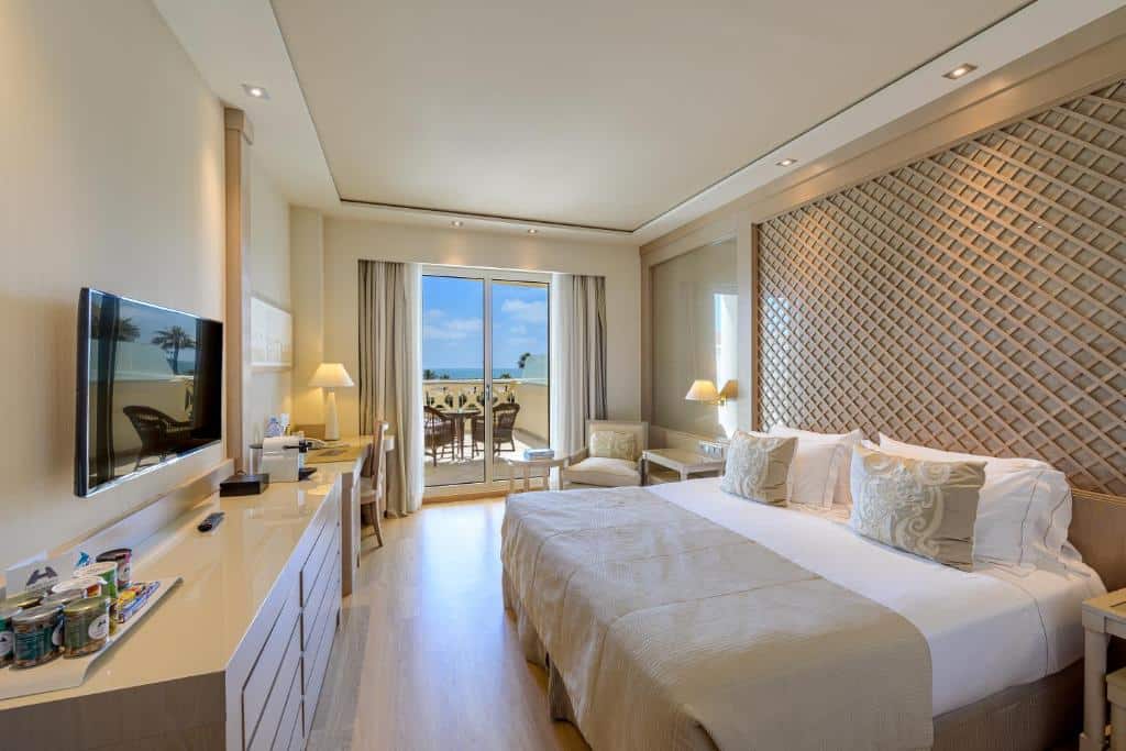 Amplo quarto do Las Arenas Balneario Resort, com cama de casal, móveis claros, televisão na parede e grandes janelas com vista para a zona exterior com palmeiras.