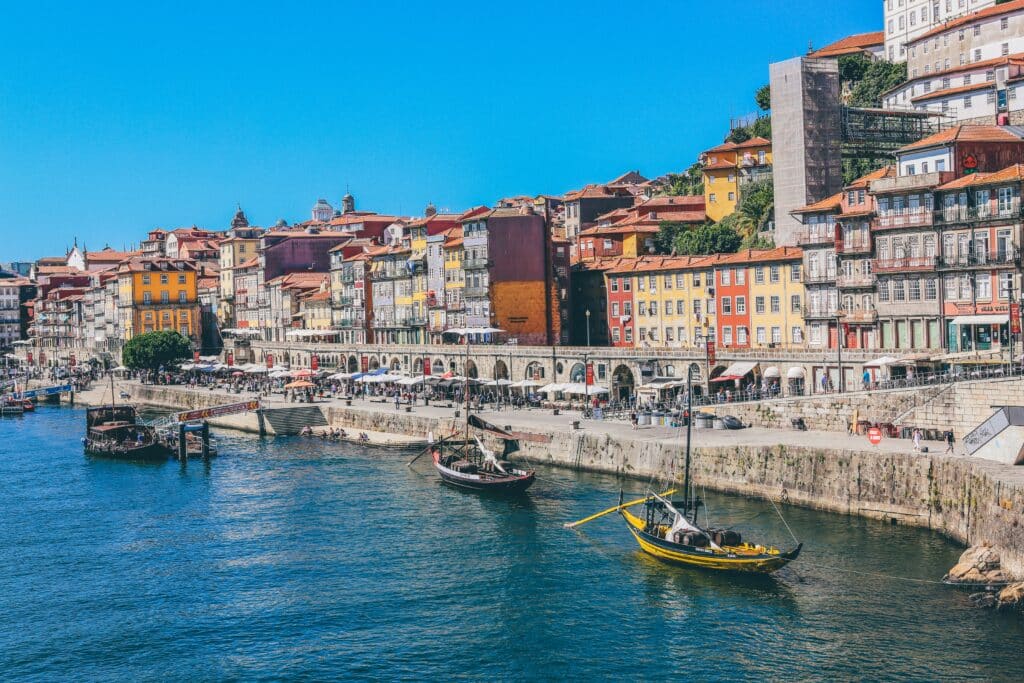 Vista do Cais da Ribeira no Porto, com barcos no rio Douro e casas coloridas ao fundo.