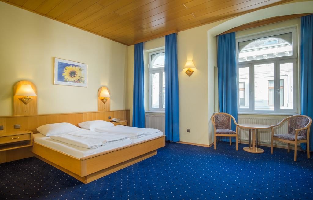 Quarto no Hotel-Pension Wild, com cama de casal, duas janelas, cortinados azuis, duas cadeiras, uma pequena mesa e apliques de parede.