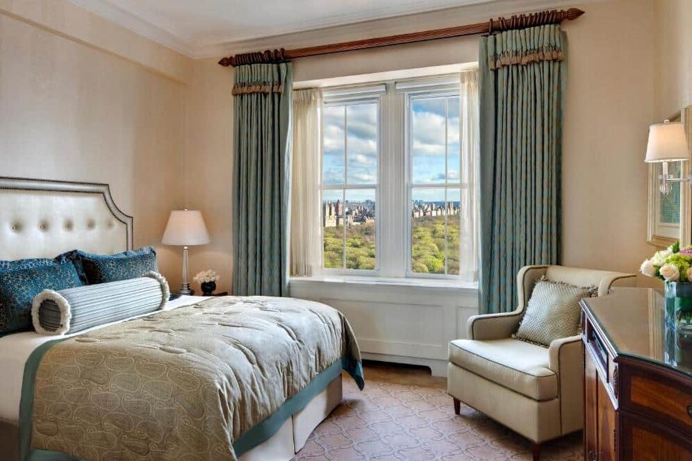 No quarto do The Pierre há uma cama de casal com cabeceira estofada branca ao centro. À direita, uma poltrona bege e uma mesa de cabeceira com candeeiro; à esquerda, um aparador com um jarro de flores. Ao fundo, uma janela ampla com cortinados verde-azulados e vista para o parque.