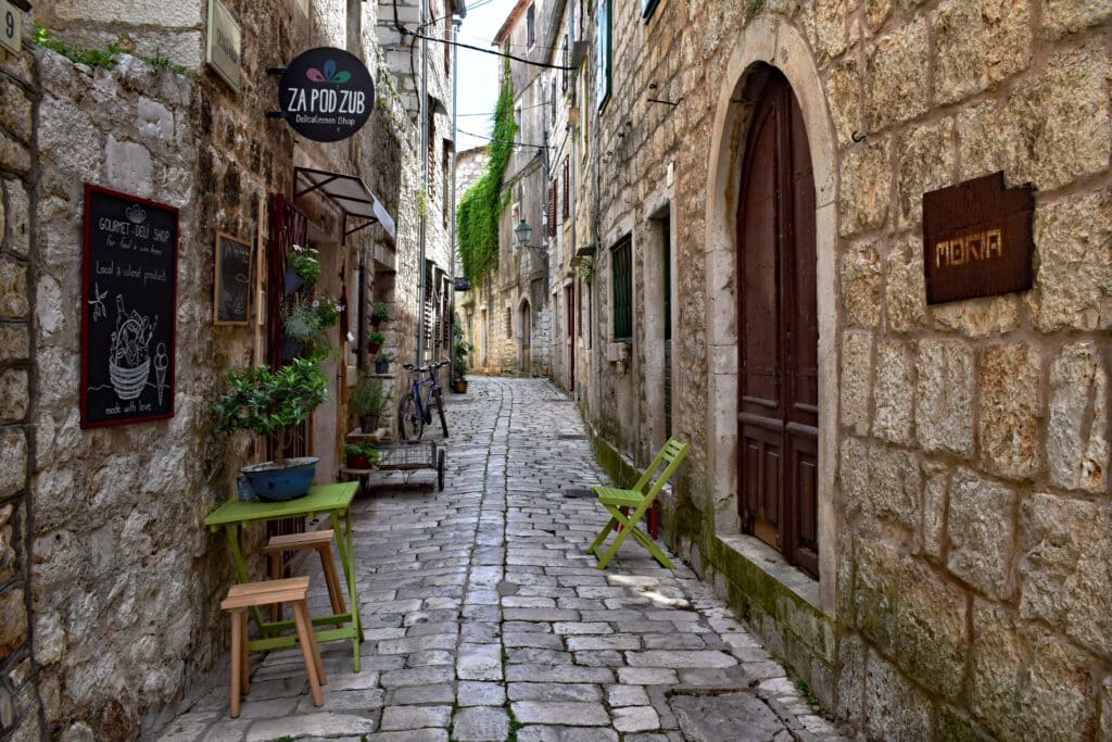 Beco estreito de calçada com um pequeno café ao ar livre em Stari Grad, com cadeiras verdes, plantas em vasos e edifícios de pedra dos dois lados.