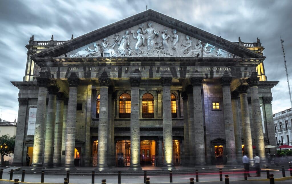 Teatro Degollado, edifício clássico de fachada ornamentada com estátuas, colunas e relevos em pedra, iluminado à noite, numa praça pública com pessoas a passar.