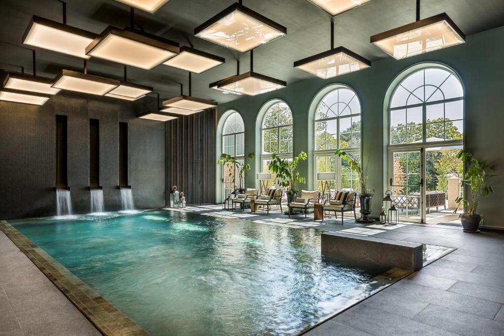 Piscina interior do Maison Albar – Amoure, com fontes de água, espreguiçadeiras, grandes janelas em arco e vasos com plantas.