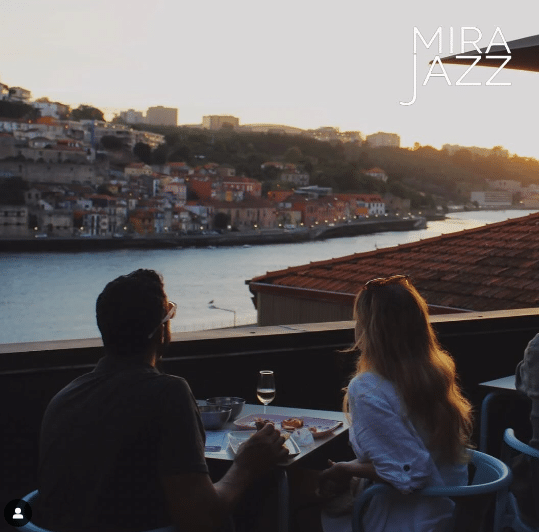 Duas pessoas a jantar num restaurante na cobertura, com vista para o rio e a cidade ao pôr do sol.