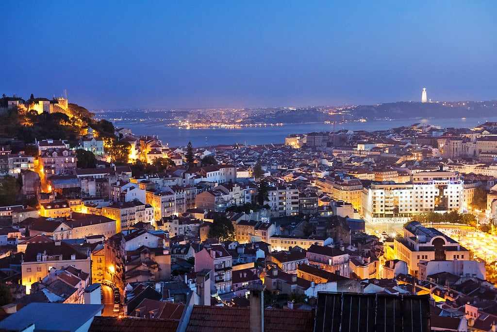 Vista do Miradouro da Senhora do Monte, em Lisboa, com parte do bairro histórico, a ponta do Castelo de São Jorge à esquerda e, ao fundo, o rio Tejo iluminado à noite