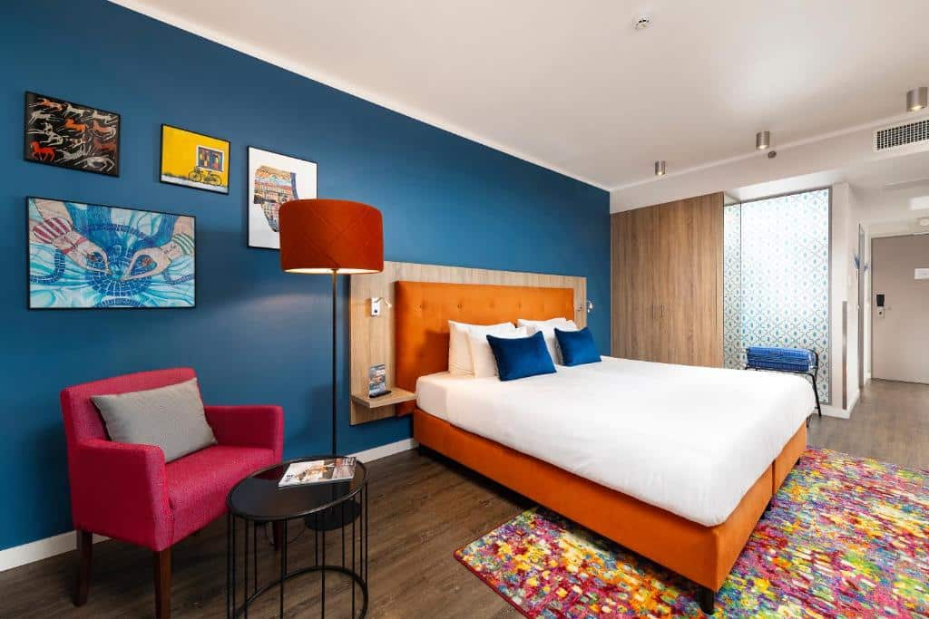 Um quarto do Three Corners Lifestyle Hotel com uma cama laranja, parede de destaque azul, cadeira vermelha, obras de arte e tapete colorido sobre o soalho.