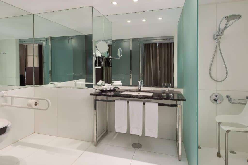 Casa de banho adaptada do Sheraton Lisboa Hotel & Spa, com lavatório baixo sem armário por baixo, toalhas em local de fácil acesso, uma cadeira dentro do duche sem ressalto e barras de apoio em redor da sanita