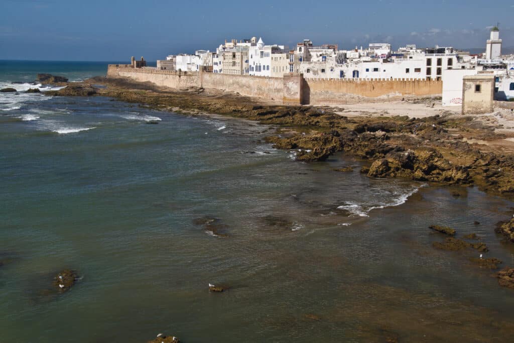 Vista costeira de Essaouira, com edifícios brancos e uma muralha ao longo da costa rochosa, virada para o oceano.
