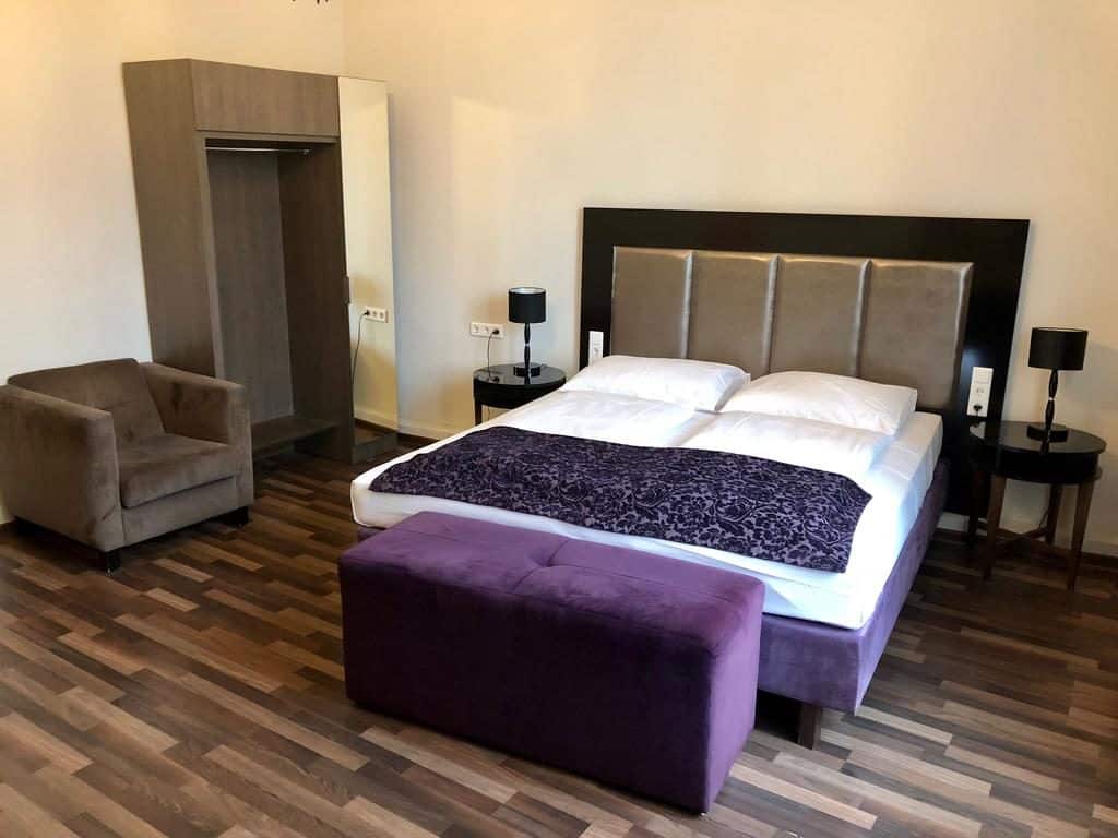 Quarto no Hotel Pension Baron am Schottentor, com cama de casal, banco roxo, poltrona, roupeiro e duas mesas de cabeceira com candeeiros.