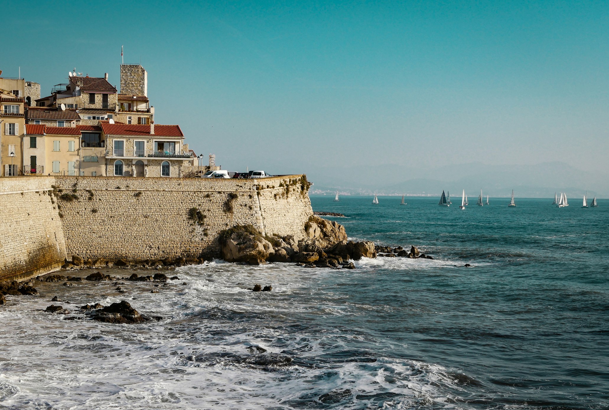 Muralha de fortaleza de pedra num litoral rochoso, com veleiros ao longe, em Antibes.
