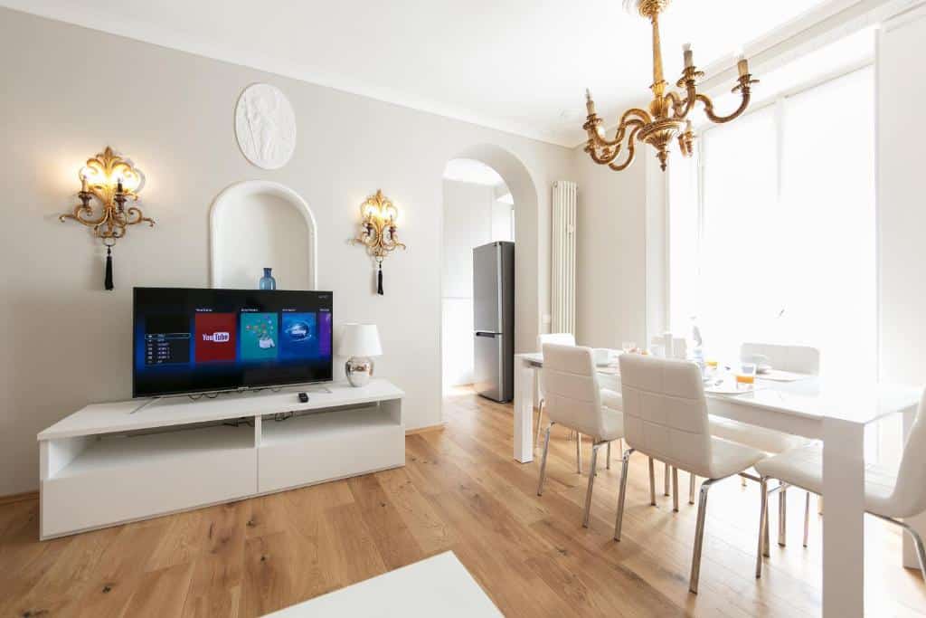 Sala de estar do Milan Retreats Duomo com um móvel de TV branco com uma televisão e peças de decoração, ao lado uma mesa de seis lugares com um lustre dourado no teto