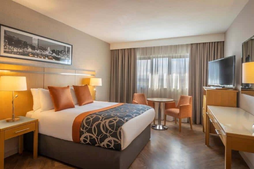 Quarto do Clayton Hotel Ballsbridge com cama de casal, decoração elegante em tons de laranja e bege, secretária e cadeiras.