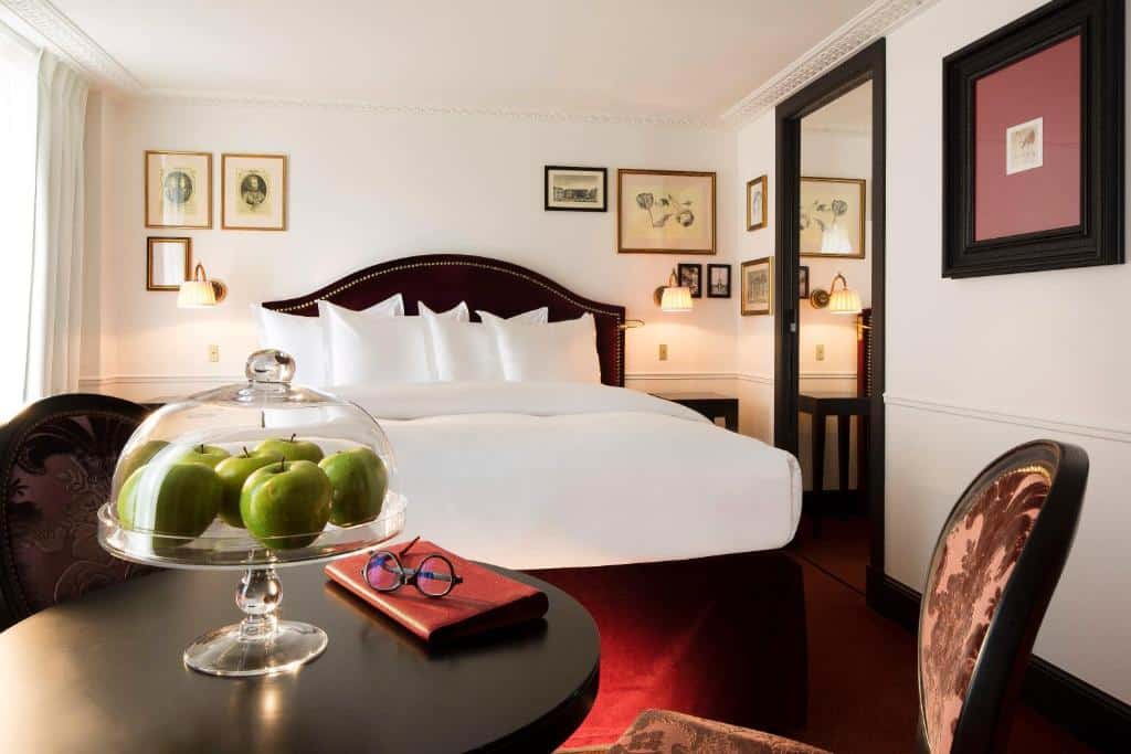 Cama bem composta num quarto luminoso do La Chambre du Marais, com arte emoldurada, uma mesa e maçãs verdes.