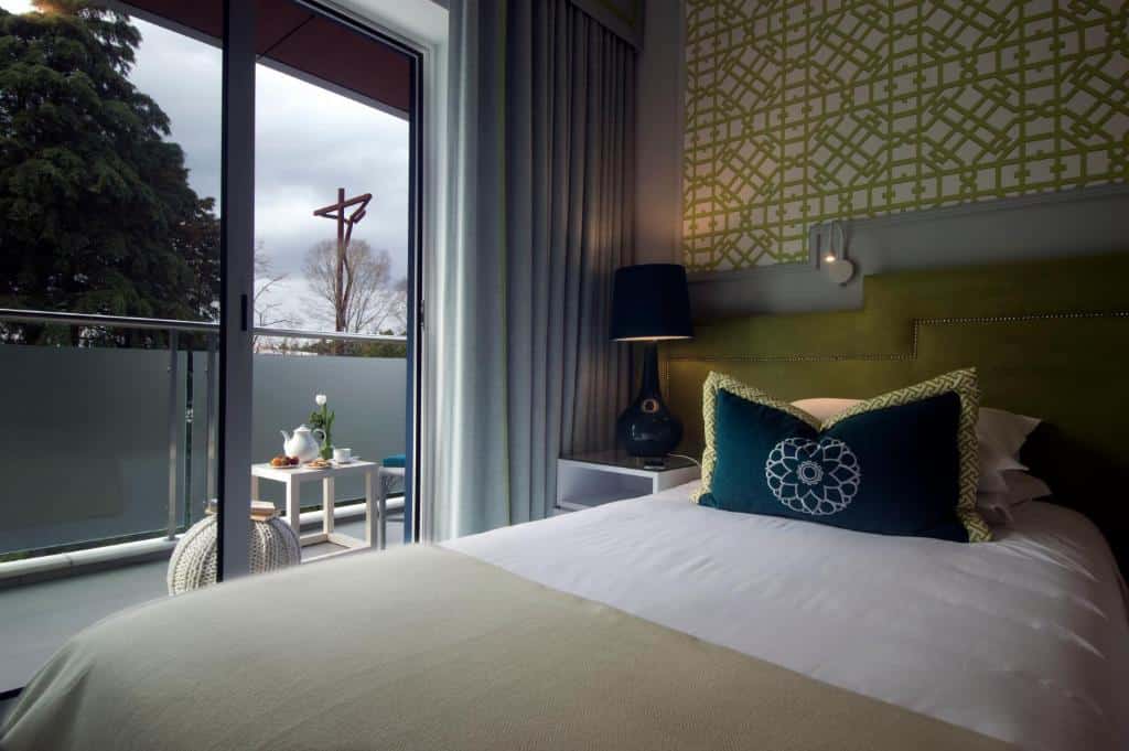 Um quarto no Coração de Fátima Boutique Hotel com uma cama, candeeiro e almofadas, ao lado de uma varanda com cadeiras e uma pequena mesa no exterior, para representar onde ficar em Fátima.