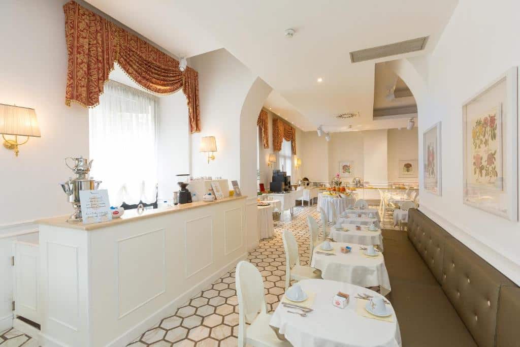 salão do Hotel Milton Roma com mesas redondas e cadeiras brancas, espaço para o pequeno-almoço, chão decorado e cortinados em tons de laranja de estilo clássico