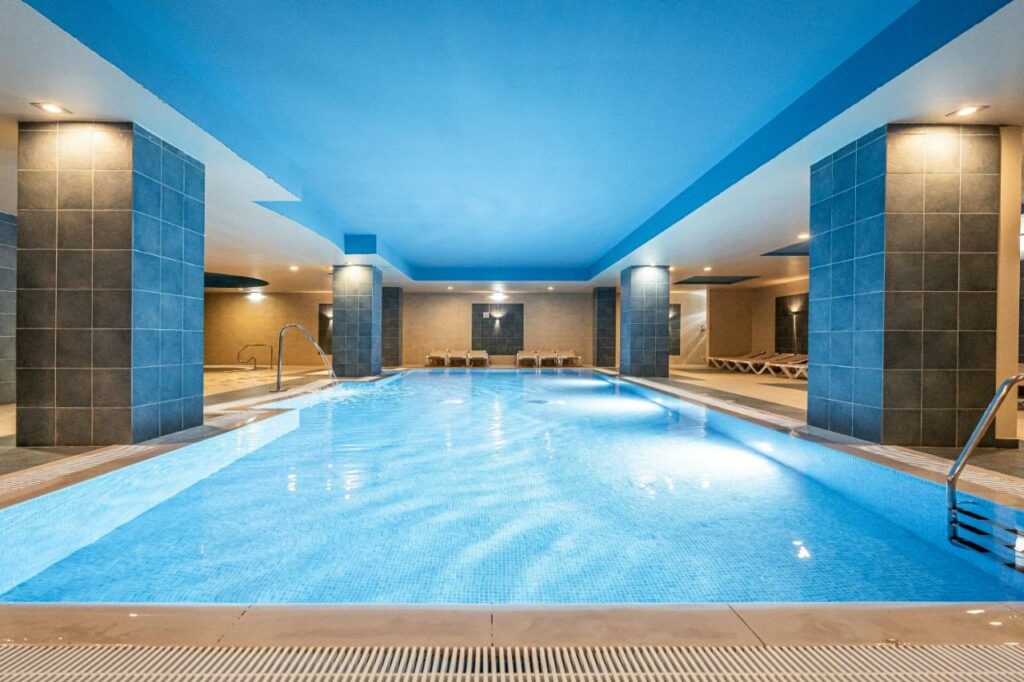Piscina coberta do 3HB Guarana – All Inclusive, sendo ela iluminada, com uma cascata pequena no meio e espreguiçadeiras em volta