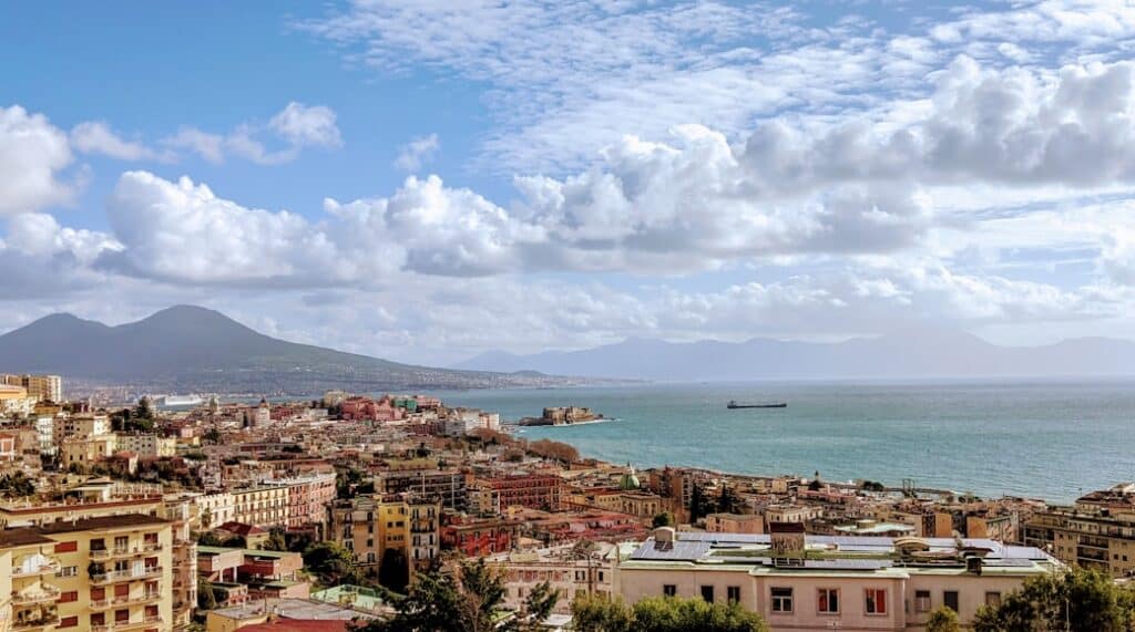 Vista panorâmica da baía de Nápoles, Itália, com montanhas ao fundo e edifícios coloridos na costa.