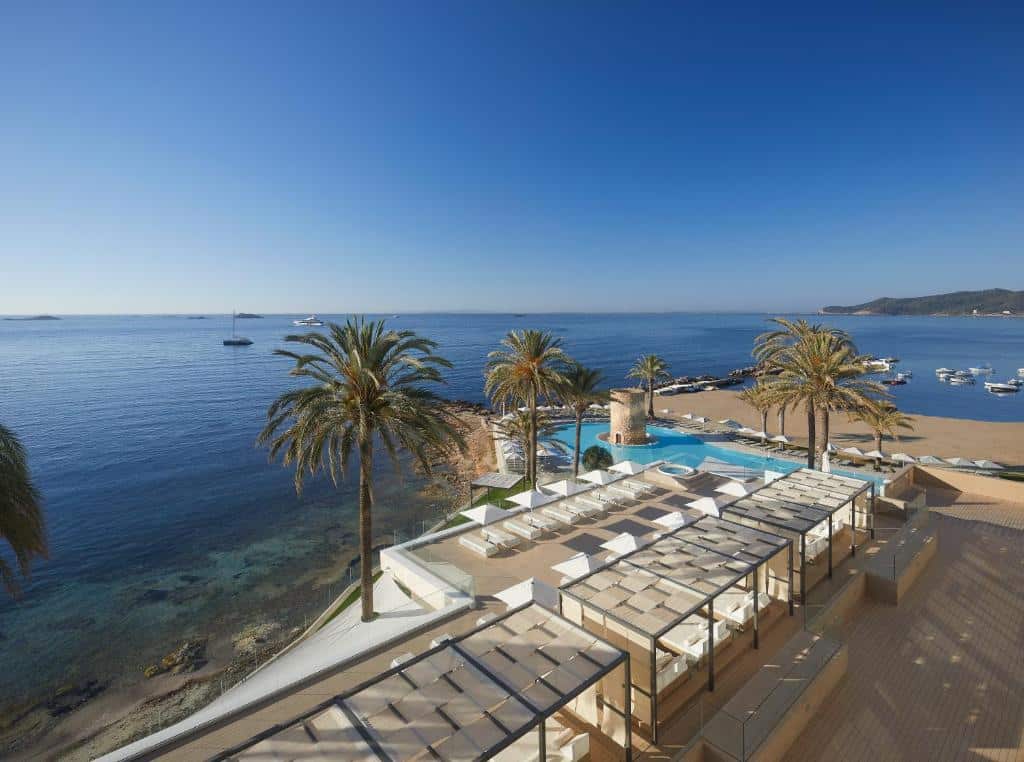 Hotel Torre del Mar à beira-mar com palmeiras, piscina, zonas de lounge e barcos ancorados em águas calmas e azuis.