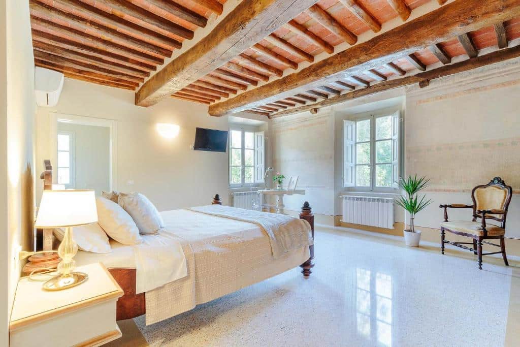 Quarto do Antica Pergola di San Giusto, um dos hotéis na Toscana, com teto de madeira, duas janelas, poltrona de época, pequena mesa redonda com um vaso de flores, cama de casal e mesa de cabeceira com candeeiro