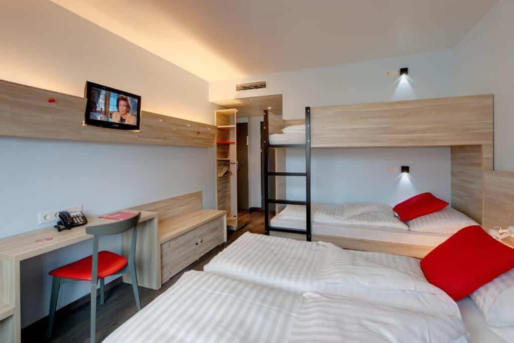 Quarto no Meininger Hotel Wien Downtown, com um beliche, uma cama de solteiro, uma secretária, uma TV e almofadas vermelhas.