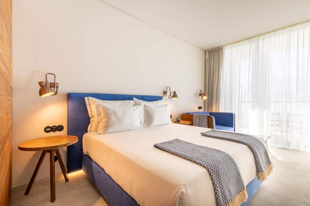 Quarto moderno no 3HB Faro, em Faro, com uma cama estofada azul, roupa de cama branca, duas mantas cinzentas e grandes cortinas transparentes que deixam entrar a luz.