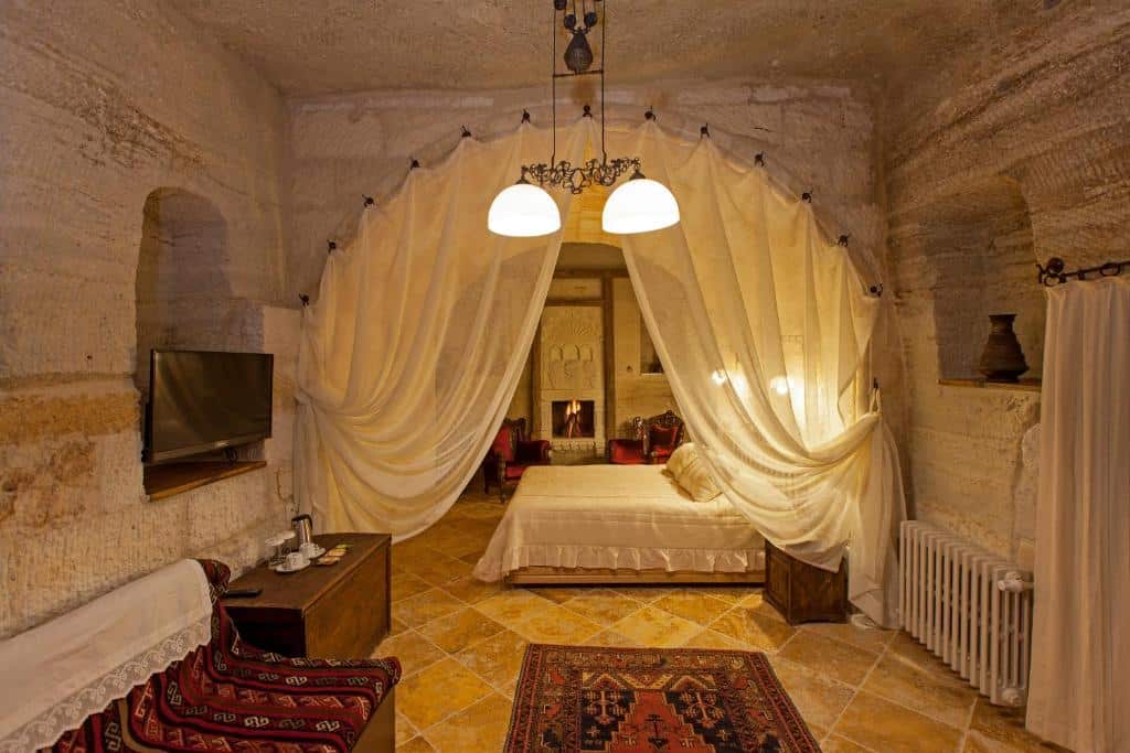 Quarto acolhedor do Mithra Cave Hotel, um dos hotéis da Capadócia, com paredes de pedra, cama atrás de cortinados drapeados, alcatifa, televisão e lareira ao fundo.