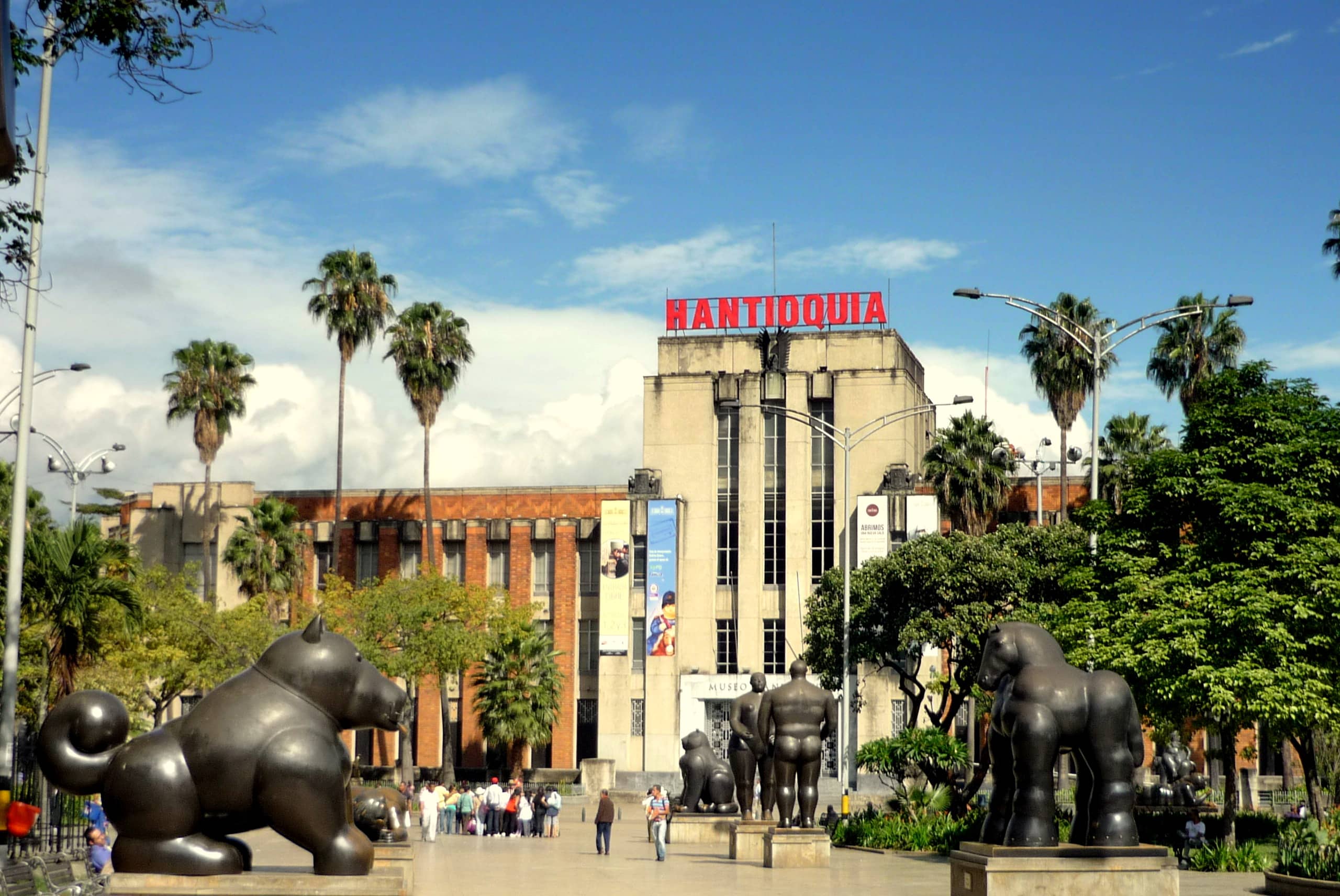 Plaza Botero, com grandes esculturas de animais, palmeiras e um edifício com a inscrição ANTIOQUIA; pessoas a passear sob um céu limpo.
