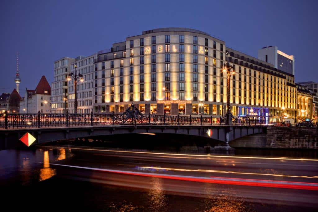Edifício iluminado do hotel Meliá Berlim, numa esquina, ao anoitecer, com uma ponte e um rio em frente.