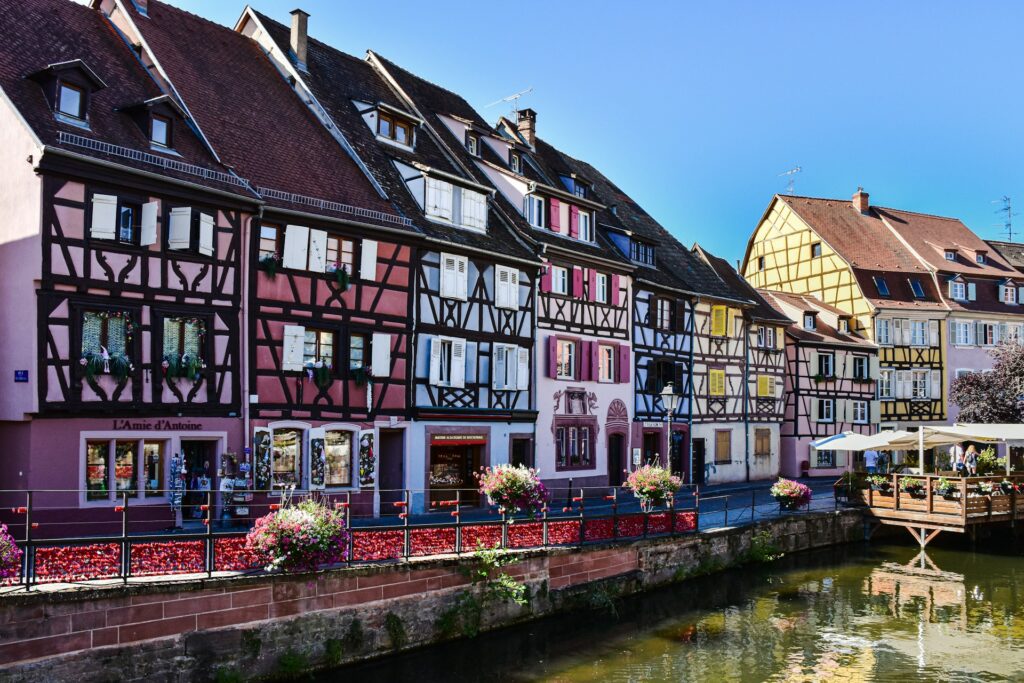 Casas coloridas em enxaimel à beira de um canal em Colmar, França. Cada edifício tem uma fachada única e janelas adornadas com floreiras cheias de flores. Um pequeno deck de madeira estende-se sobre o canal, que reflete as cores das casas e das flores.