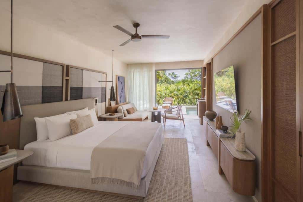 Quarto do Alila Mayakoba com cama king size, decoração neutra com sofá-cama ao fundo, TV, poltronas e varanda com piscina e vista para a vegetação.