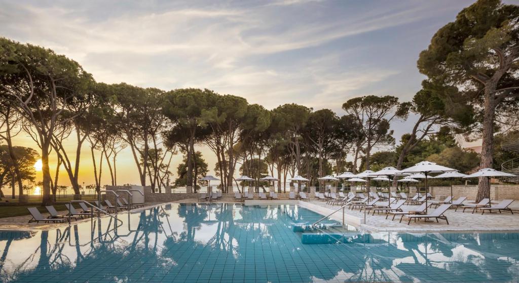 Piscina de hotel com espreguiçadeiras e chapéus de sol ao pôr do sol, rodeada de pinheiros à beira-mar, no Falkensteiner Hotel Adriana l Adults Only