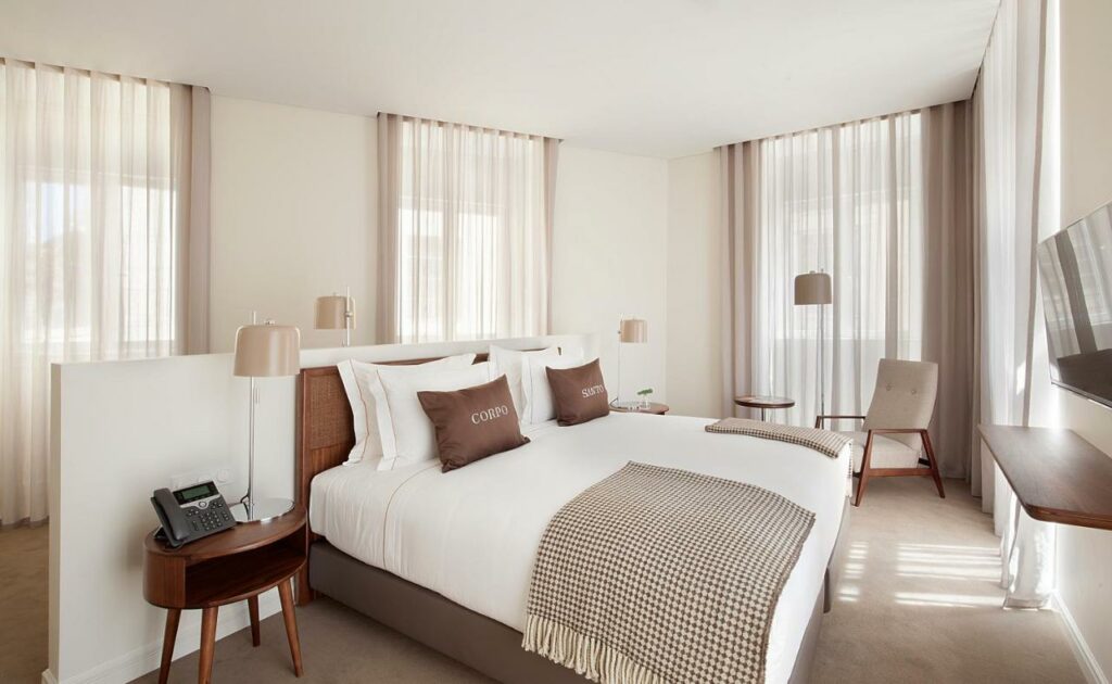 Quarto moderno no Corpo Santo Lisbon Historical Hotel com uma cama grande, decoração neutra, duas cadeiras, uma mesa lateral e uma TV na parede.