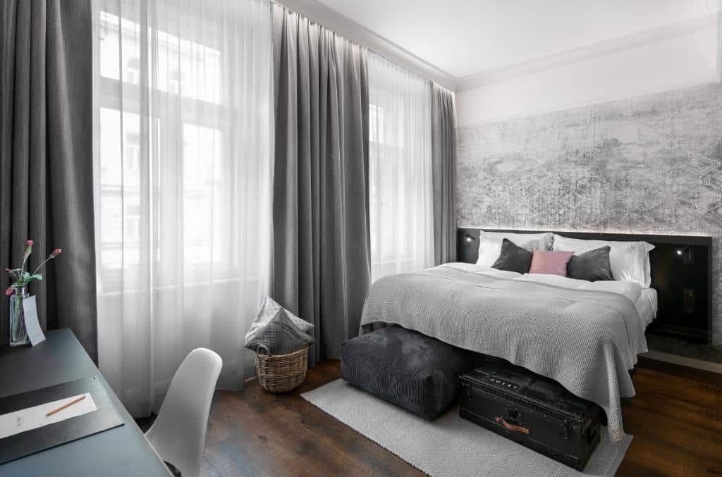 Quarto moderno do The Manes Boutique Hotel Prague, com cama de casal, roupa de cama cinzenta, janelas grandes e uma secretária preta.