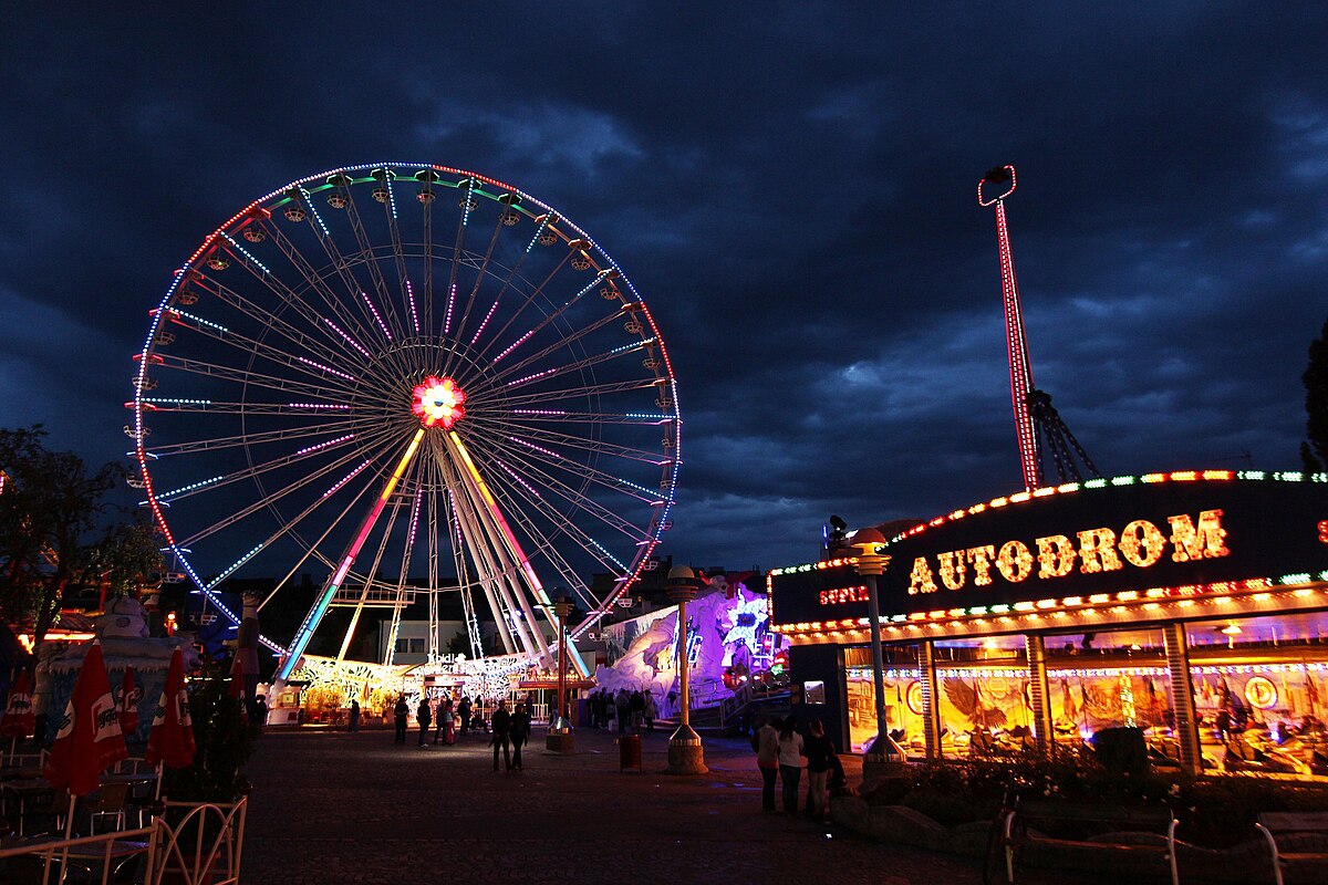 Prater à noite, com a roda gigante iluminada e pessoas a passear, sob um céu nublado.