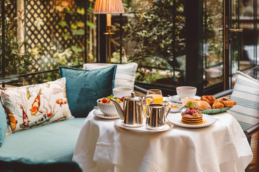 Mesa posta para o pequeno-almoço no Hôtel de l'Abbaye, com café, sumo, bolos e fruta, numa sala iluminada pelo sol.