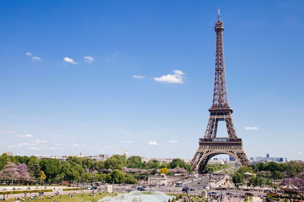 A Torre Eiffel em Paris destaca-se num céu azul, com fontes, vegetação e pessoas em primeiro plano.