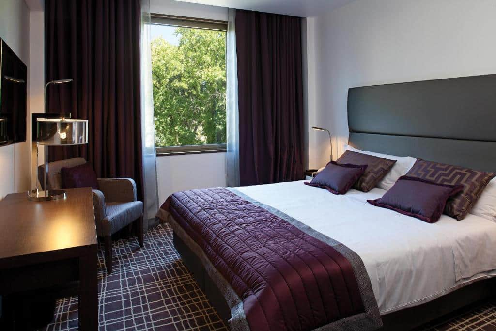 Quarto do NEYA Lisboa Hotel com uma cama de casal, uma janela com cortinas, alcatifa em tons de branco e roxo, uma mesa de madeira com televisão em frente à cama e uma poltrona junto à janela.