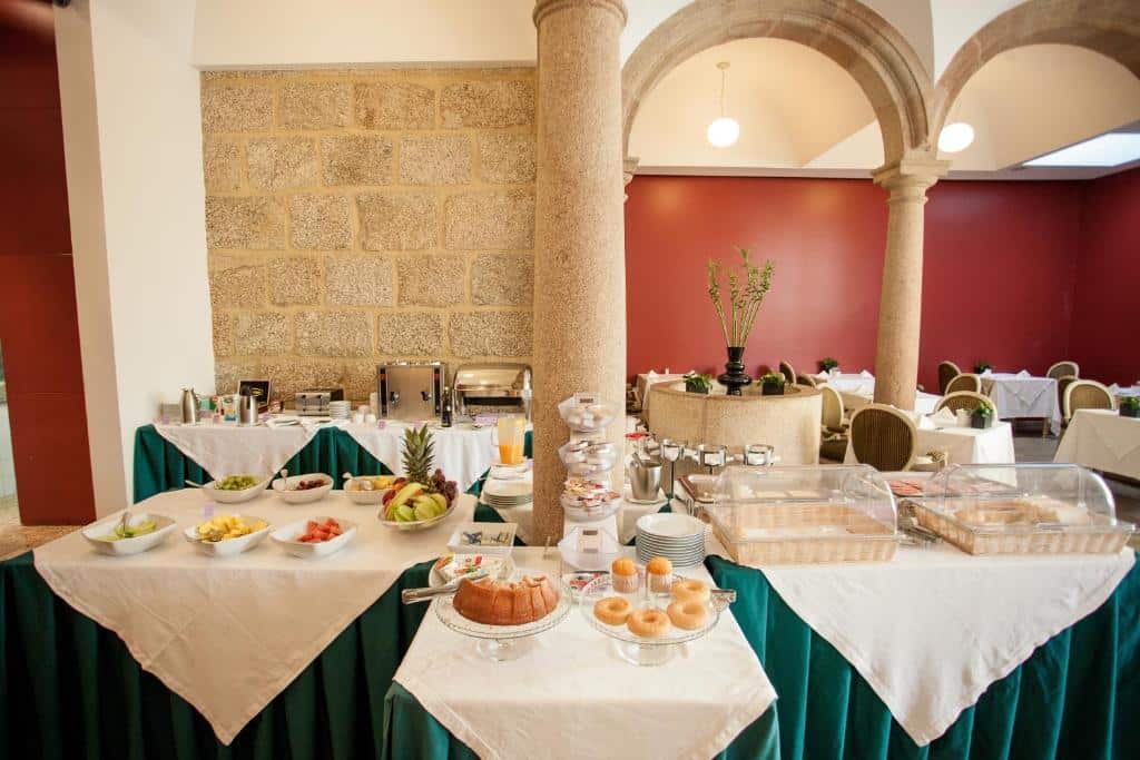 Buffet de pequeno-almoço no Hotel Bracara Augusta, com bolos, fruta e bebidas dispostos numa sala com colunas de pedra.