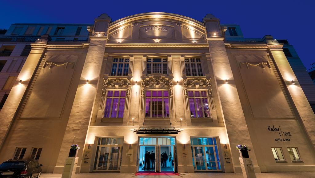 Entrada do Ruby Sofie Hotel bem iluminada à noite, com grandes janelas e pessoas visíveis no interior.