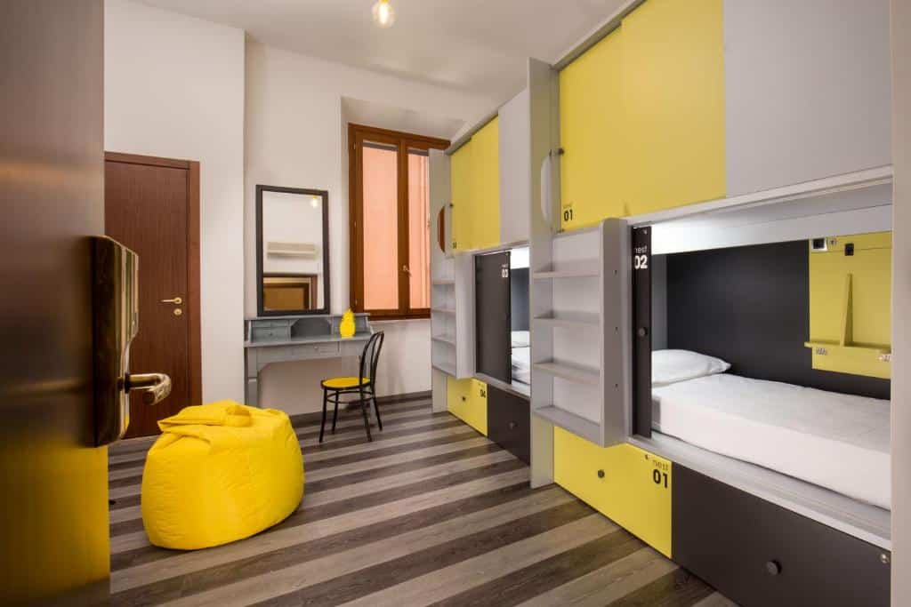 quarto do Free Hostels Roma, com mesa, cadeira e espelho, pufe amarelo e dois beliches amarelos com escada e cofre, junto a uma janela de madeira