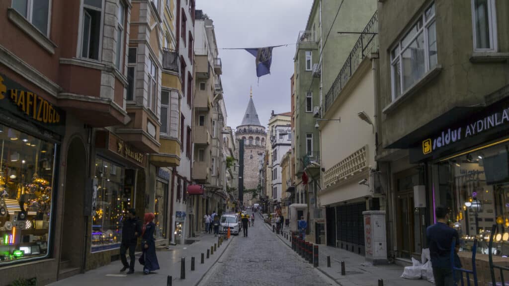 Uma rua estreita de calçada repleta de lojas e edifícios que conduz à Torre de Gálata em Istambul, sob um céu nublado.