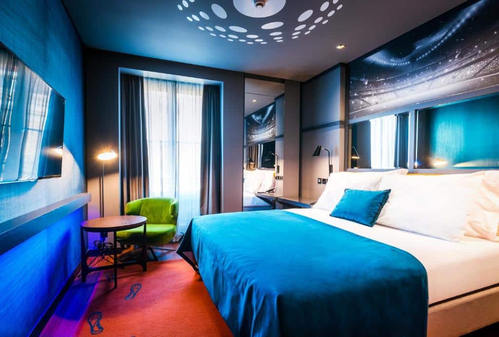 Quarto do Pestana CR7 Lisboa com decoração em tons de azul e vermelho, uma cama de casal, uma pequena varanda com cortinas, uma televisão em frente à cama e uma pequena mesa redonda junto de um cadeirão