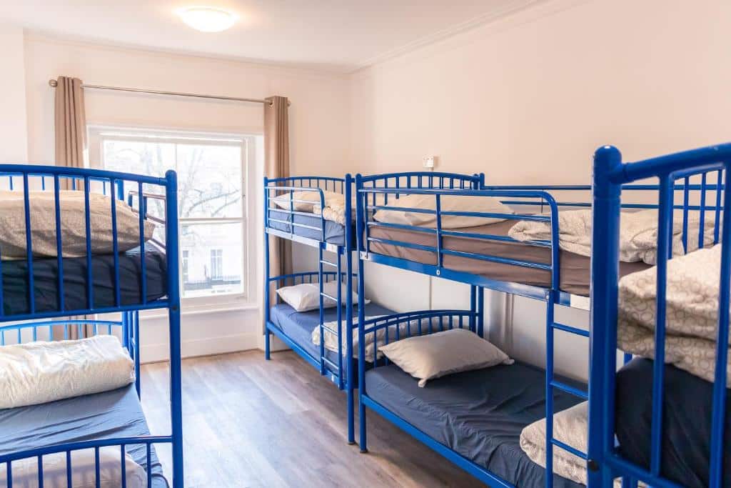 Quarto do Urbany Hostel London com vários beliches azuis com almofadas, uma janela ampla com cortina e chão de madeira.