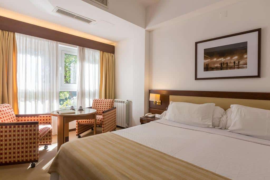 Quarto no Hotel São José com cama de casal, duas poltronas, uma pequena mesa junto à janela e um quadro emoldurado na parede.