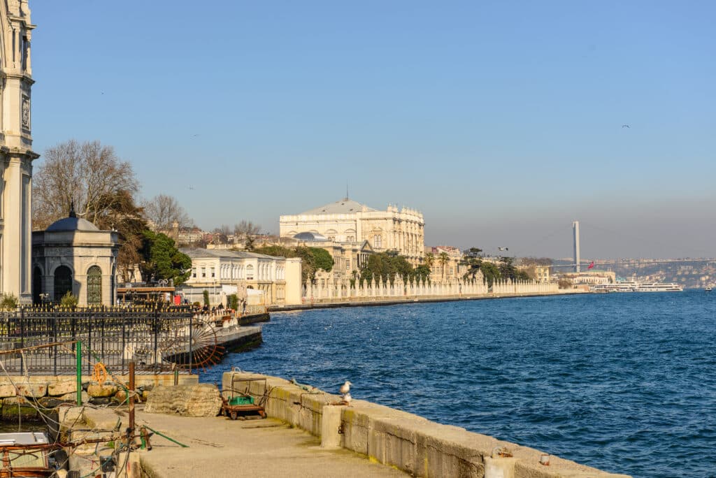 Um passeio à beira-mar com edifícios históricos, água azul calma e uma ponte suspensa ao longe, sob um céu claro.